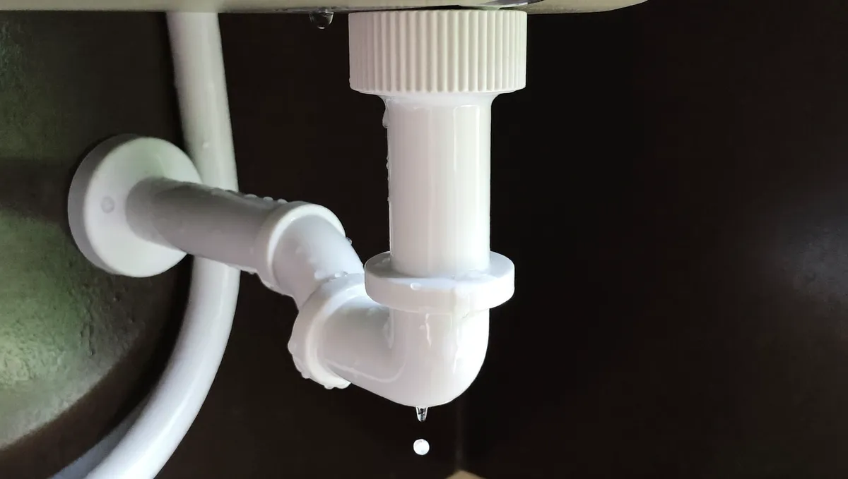 White PVC pipe joint under a sink with a water drop leaking from the connection